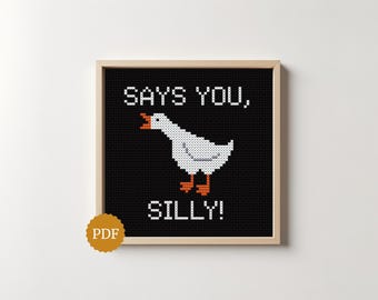 May include: A cross-stitch pattern in a light wood frame featuring a white goose with an orange beak and feet on a black background. The text SAYS YOU, SILLY! is stitched in white above and below the goose. A gold circular PDF icon is in the corner.