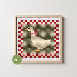 Op de afbeelding: Een vierkant kruissteekpatroon in een lichthouten lijst, met een witte gans met een oranje snavel en poten, die een rode strik draagt. De gans is gecentreerd op een donker olijfgroene achtergrond, omringd door een rood-witte geblokte rand.