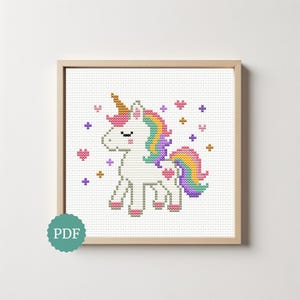 May include: A framed cross-stitch pattern of a white unicorn with a rainbow mane and tail, a golden horn, and pink accents. The design includes pink hearts and purple stars. The frame is a light brown color. The image has a PDF icon.