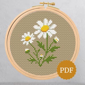 May include: Cross-stitch embroidery of daisies in a wooden hoop. The daisies have white petals and yellow centers, with green leaves and stems. The background is a tan fabric. A PDF icon is in the lower right corner.