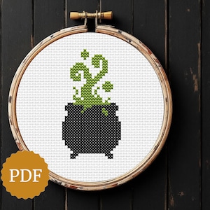 May include: Cross-stitch design of a black cauldron with green bubbling liquid and a swirling green design above it, set in a wooden embroidery hoop. The image has a PDF logo in the lower left corner.