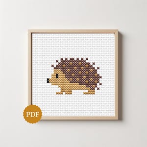 Könnte beinhalten: Ein Kreuzstichmuster mit einem kleinen Igel, der einen dunkelbraunen Rücken sowie ein hellbraunes Gesicht, einen Körper und Füße hat. Das Design ist auf einem weißen Rasterhintergrund zentriert und in einem hellen Holzrahmen gerahmt. Ein goldenes PDF-Abzeichen ist zu sehen.