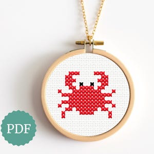 May include: A cross-stitch crab design in red and black is displayed within a wooden embroidery hoop, suspended by a gold-colored chain. The crab is centered on a white fabric background. A teal-colored PDF icon is in the lower left.