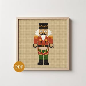 May include: A cross-stitch pattern of a nutcracker soldier wearing a red jacket with gold accents, a black hat, and green trousers. The design is displayed in a light wood frame on a tan Aida cloth background with a circular PDF icon in the corner.