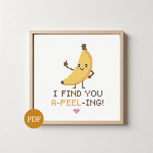 Può includere: Un motivo a punto croce in una cornice di legno chiaro raffigurante una banana gialla con un volto sorridente, braccia e gambe. Il testo recita I FIND YOU A-PEEL-ING! in lettere marroni e arancioni, con una piccola icona a cuore rosa sotto.