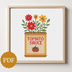 May include: Cross-stitch art of a tomato sauce can with red, yellow, and green flowers. The can has the words "TOMATO SAUCE" and a tomato graphic. The artwork is framed in a light brown wooden frame.