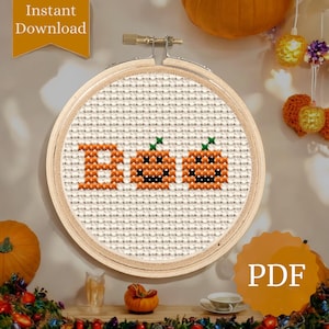 May include: A cross-stitch embroidery hoop featuring the word "Boo" and two jack-o'-lanterns. The design is stitched in orange and black thread on a cream-colored fabric. The wooden hoop is displayed against a wall with pumpkins and decorative lights.