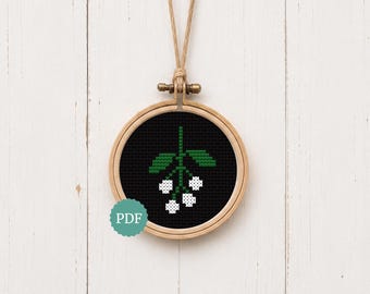 May include: A cross-stitch pattern displayed in a round wooden embroidery hoop. The design features a black background with a green stem and leaves, and four white flower clusters. A small teal circular icon with the text PDF is visible.