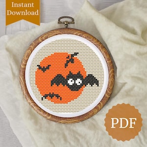 May include: A cross-stitch embroidery in a wooden hoop features a Halloween design. An orange moon is the backdrop for a black bat with white eyes. The design includes the words "Instant Download" and "PDF".