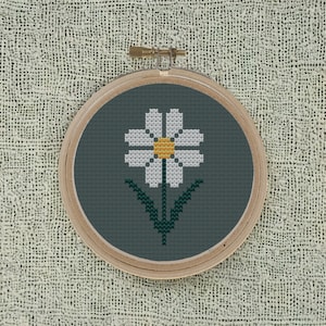 May include: A white daisy with a yellow center is embroidered on a dark green fabric in a wooden embroidery hoop.