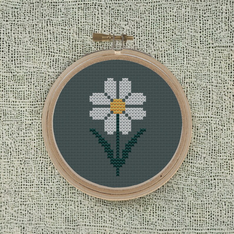 May include: A white daisy with a yellow center is embroidered on a dark green fabric in a wooden embroidery hoop.