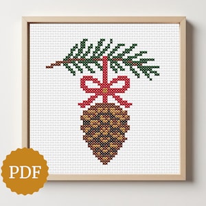 May include: Cross-stitch image of a pinecone suspended from a green branch with a red bow. The artwork is framed in a light wood frame and set against a white background. A PDF logo is in the lower left corner.