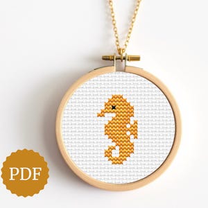 Può includere: Una collana color oro con un piccolo ciondolo rotondo in legno ricamato. All'interno del cerchio c'è un disegno a punto croce di un cavalluccio marino giallo. La collana ha una catena e una chiusura dorate. Un'icona PDF si trova in basso a sinistra.