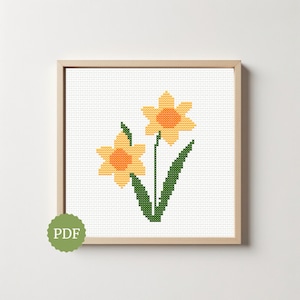 May include: A framed cross-stitch pattern of two yellow daffodils with orange centers and green stems and leaves. The artwork is in a light wooden frame and has a PDF logo in the lower left corner.