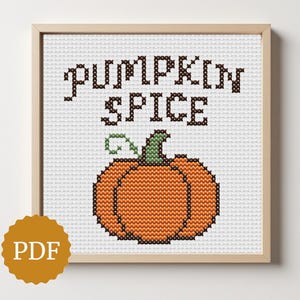 May include: A framed cross-stitch design featuring the words "PUMPKIN SPICE" above an orange pumpkin with a green stem. The artwork is in a wooden frame and is set against a white background. A PDF icon is in the lower left corner.