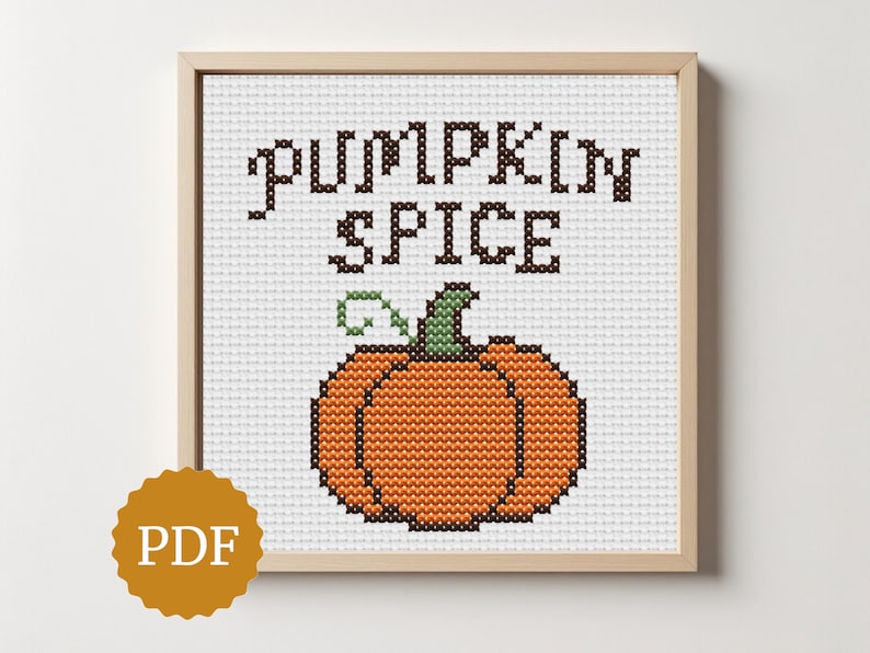 May include: A framed cross-stitch design featuring the words "PUMPKIN SPICE" above an orange pumpkin with a green stem. The artwork is in a wooden frame and is set against a white background. A PDF icon is in the lower left corner.