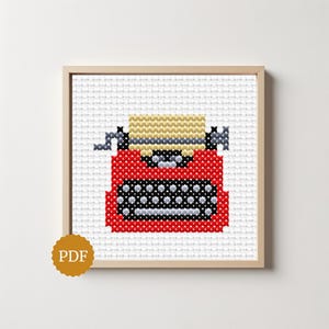 Op de afbeelding: Een kruissteekpatroon met een rode typemachine met een beige papierwagen, zwarte toetsen en grijze accenten. Het ontwerp wordt weergegeven in een lichthouten lijst tegen een witte achtergrond, met een rond goudkleurig PDF-pictogram.