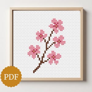 May include: Cross-stitch artwork of a cherry blossom branch with pink flowers and a brown stem, set against a white background. The artwork is framed in a light wood frame. A PDF icon is in the lower left corner.