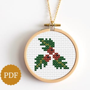 Puede incluir: Un collar dorado con un colgante con un diseño de punto de cruz de hojas y bayas de acebo. El diseño está dentro de un pequeño bastidor de bordado de madera redondo. El colgante cuelga de una delicada cadena dorada. Un icono PDF está en la parte inferior izquierda.