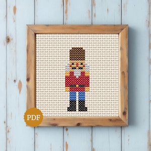 Può includere: Un motivo a punto croce di uno schiaccianoci con cappello marrone, giacca rossa con cintura dorata, pantaloni blu e stivali neri, centrato su un tessuto color crema e racchiuso in una cornice quadrata di legno.