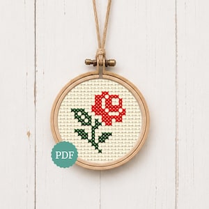 May include: A circular wooden embroidery hoop containing a cross-stitch pattern of a single red rose with green leaves on a cream-coloured fabric background. A light brown string is attached to the top of the hoop for hanging. A teal PDF icon is visible.
