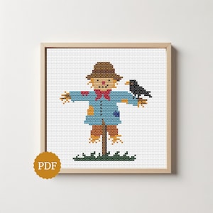 May include: A cross-stitch pattern featuring a scarecrow wearing a brown hat, a blue jacket with patches, and a red bowtie, standing on a green grassy patch with a black crow perched on its arm, all framed in a light wood square frame with a PDF icon.