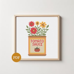 May include: A cross-stitch pattern in a light wood frame featuring a yellow tomato sauce can with red text and a small tomato graphic. Above the can are red and yellow flowers with green leaves on a white grid background. A gold PDF icon is visible.