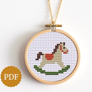 May include: A gold-colored necklace featuring a small, round pendant with a cross-stitch design of a rocking horse. The horse is beige, brown, red, and green, set against a white background within a wooden hoop.