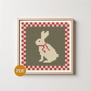 May include: A cross-stitch pattern in a light wood frame featuring a white rabbit with a red scarf centred on a dark olive green background. The design is enclosed by a red and white chequered border. A gold circular icon with PDF text is visible.
