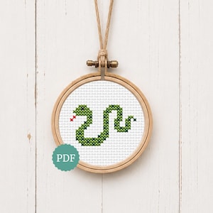 May include: A circular wooden embroidery hoop containing white fabric with a cross-stitch pattern of a green snake with a red tongue. A teal circular icon with the white text PDF is positioned in the lower left corner of the embroidery hoop.