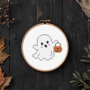 May include: A circular wooden embroidery hoop containing a cross-stitch pattern of a white ghost with black eyes, a black mouth, and pink cheeks. The ghost is holding an orange jack-o'-lantern bucket with a black face design on its handle.