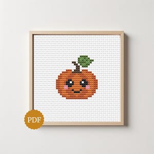 May include: A cross-stitch pattern of a small orange pumpkin with a green leaf, a brown stem, and a smiling face with black eyes and pink cheeks, displayed in a light wood frame with a gold circular PDF icon in the bottom left corner.
