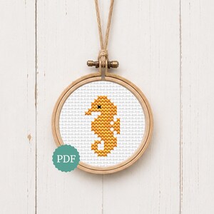 May include: A circular wooden embroidery hoop containing a white fabric panel with a cross-stitched orange seahorse design. The hoop is suspended by a tan twine loop at the top. A small teal circular icon with the white text PDF is visible.