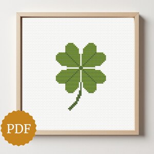 Puede incluir: Un bordado a punto de cruz enmarcado de un trébol de cuatro hojas en tonos verdes. La obra de arte está sobre un fondo blanco y enmarcada en madera clara. Un icono PDF dorado está en la esquina inferior izquierda.