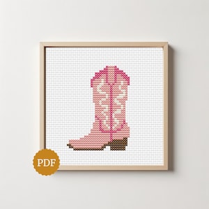 May include: A cross-stitch pattern featuring a pink cowboy boot with white decorative stitching and a brown heel, displayed in a light wood frame. A circular gold badge with the text PDF is positioned in the lower left corner of the frame.