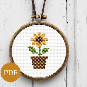 Könnte beinhalten: Ein Kreuzstich-Anhänger mit einer Sonnenblume in einem braunen Topf. Die Blume hat gelbe Blütenblätter und eine braune Mitte, mit grünen Blättern und Stiel. Der Anhänger befindet sich in einem runden, bronzefarbenen Rahmen mit einer braunen Lederschnur. Das Bild enthält auch ein PDF-Logo.