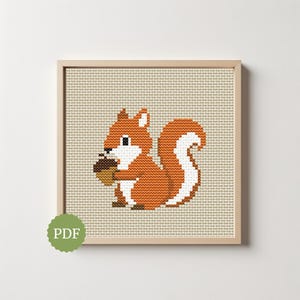 Op de afbeelding: Kruissteekkunst van een gepixeleerd eekhoorntje met een eikel. Het eekhoorntje is oranje en wit, tegen een beige achtergrond. Het kunstwerk is ingelijst in een lichtbruine houten lijst. Een groen PDF-pictogram staat linksonder.