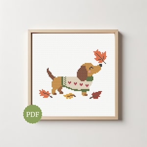 May include: A cross-stitch pattern featuring a brown dachshund wearing a white jumper with red hearts and green trim. The dog is surrounded by scattered orange, yellow, and dark red autumn leaves. The design is displayed in a light wood frame.