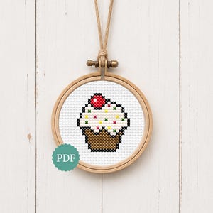 May include: A cross-stitch pattern of a cupcake with white frosting, colourful sprinkles, and a red cherry on top, displayed in a round wooden embroidery hoop. The design features a brown cupcake liner and includes a circular teal PDF label.