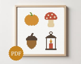 May include: Cross-stitch art print featuring autumn-themed designs: a pumpkin, mushroom, acorn, and lantern with a candle. The artwork is framed in a light wood frame and includes a PDF icon in the lower left corner. The designs are in shades of orange, brown, and red.