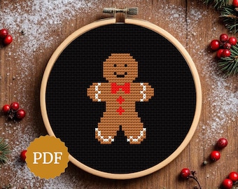 May include: A cross-stitch design of a gingerbread man with a red bow tie and white icing details, set against a black background within a wooden embroidery hoop. The PDF icon is visible. Festive holiday decor.