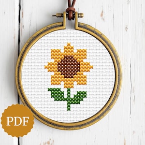 Può includere: Un disegno di girasole a punto croce in un telaio rotondo color oro antico. Il girasole ha petali gialli, un centro marrone e foglie e stelo verdi. Il telaio è sospeso da un cordino in pelle marrone. Un logo PDF è in basso a sinistra.