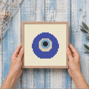 May include: A square wooden frame containing a cross-stitch embroidery of a nazar amulet. The design features a central black pupil surrounded by concentric circles of light blue, white, and a large outer ring of dark blue on a cream background.