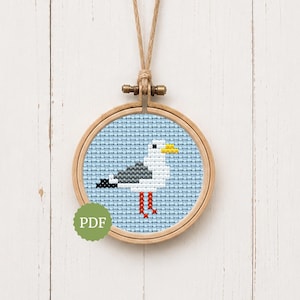 Puede incluir: Un patrón de punto de cruz que muestra una gaviota pixelada con plumas blancas, grises y negras, un pico amarillo y patas naranjas sobre un fondo azul claro, presentado en un bastidor de madera con cuerda para colgar y una etiqueta PDF verde.