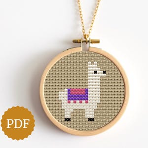 May include: A gold-colored chain necklace with a round pendant featuring a cross-stitch llama design. The llama is white with a pink and purple blanket. The pendant is set in a wooden embroidery hoop.
