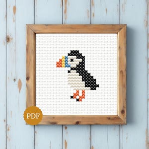 Puede incluir: Un patrón de punto de cruz que muestra un frailecillo pixelado con espalda negra, pecho blanco y un pico multicolor en rojo, amarillo y azul, presentado en un marco de madera cuadrado con un icono PDF circular en la esquina inferior izquierda.