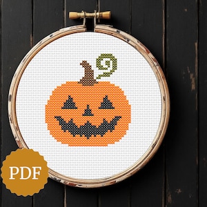 Può includere: Un disegno a punto croce di una zucca sorridente in un telaio da ricamo. La zucca è arancione con occhi e bocca neri e un gambo verde. Il telaio è posto su uno sfondo di legno scuro. Un'icona PDF è in basso a sinistra.