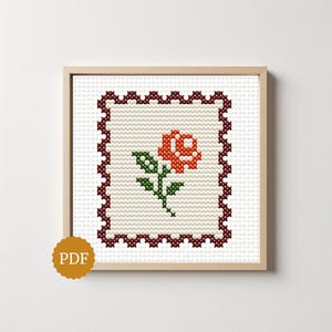 May include: A cross-stitch pattern featuring a central orange rose with green leaves on a white grid background, framed by a dark red scalloped border. A circular gold icon with the text PDF is positioned in the lower left corner of the frame.
