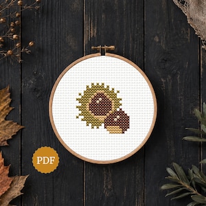 May include: A circular wooden embroidery hoop containing a white fabric panel with a cross-stitch pattern of two brown chestnuts, one partially encased in a spiky green husk, featuring a small yellow circular icon with the text PDF.