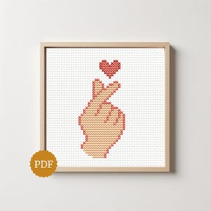 May include: A cross-stitch pattern framed in light wood featuring a hand making a finger heart gesture with a small red heart above it. The design is rendered in a pixelated style on a white grid background, with a gold circular PDF icon in the corner.